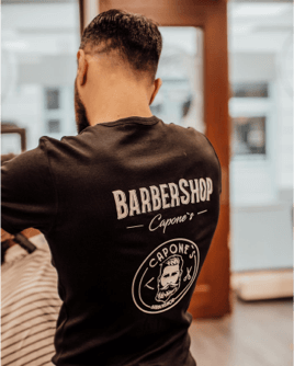 Ein Barber von Capone’s Barbershop in einem schwarzen Shirt mit dem Logo und Schriftzug des Barbershops, während er an einem Kunden arbeitet.